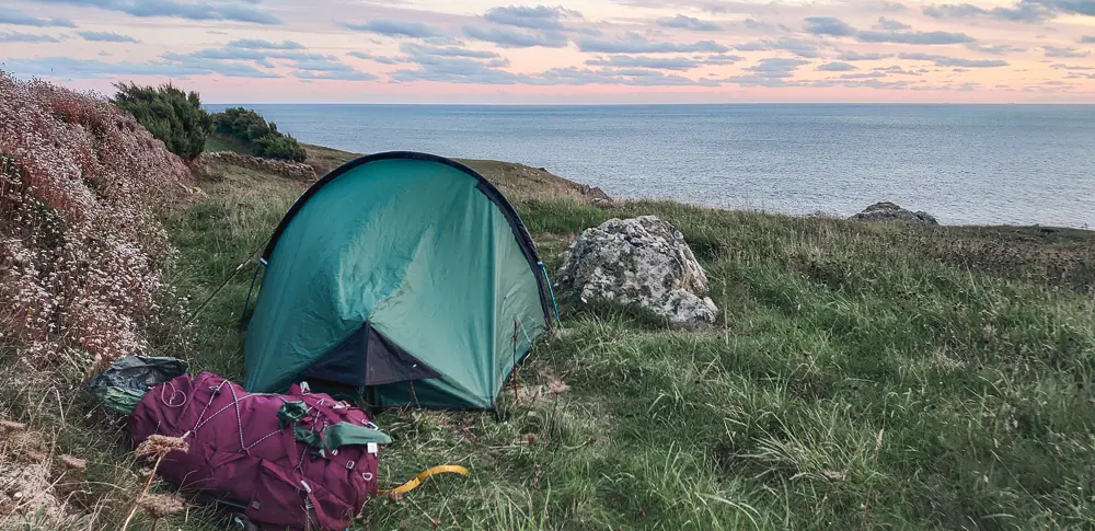 A small tent is pitched beside a stone wall on the South West Coast PAth in Cornwall. The sky is pink above a cold-looking sea.