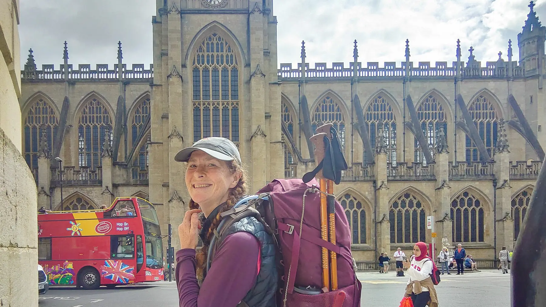 Looking over my shoulder, smiling despite the weight of my backpack, with Bath Abbey and a red bus in the background