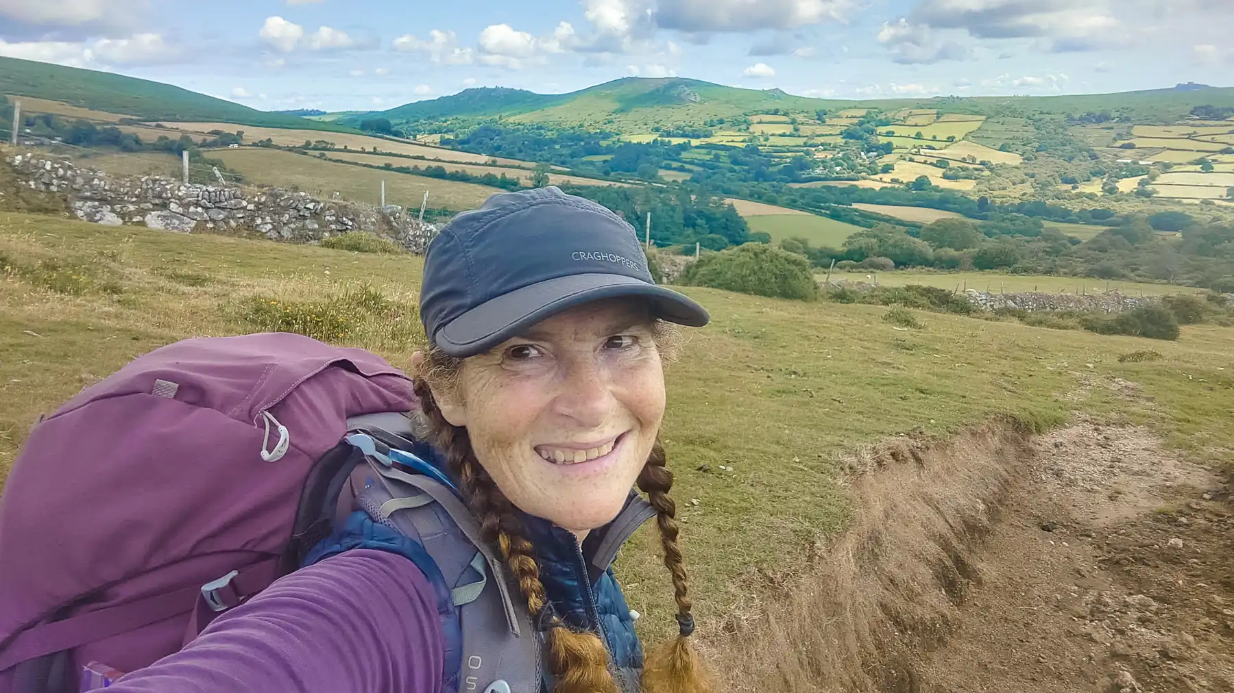 Stephanie Boon, smiling at the camera, is hiking on a sunny day on Dartmoor. On the horizon, is a patchwork of fields on rough ground.