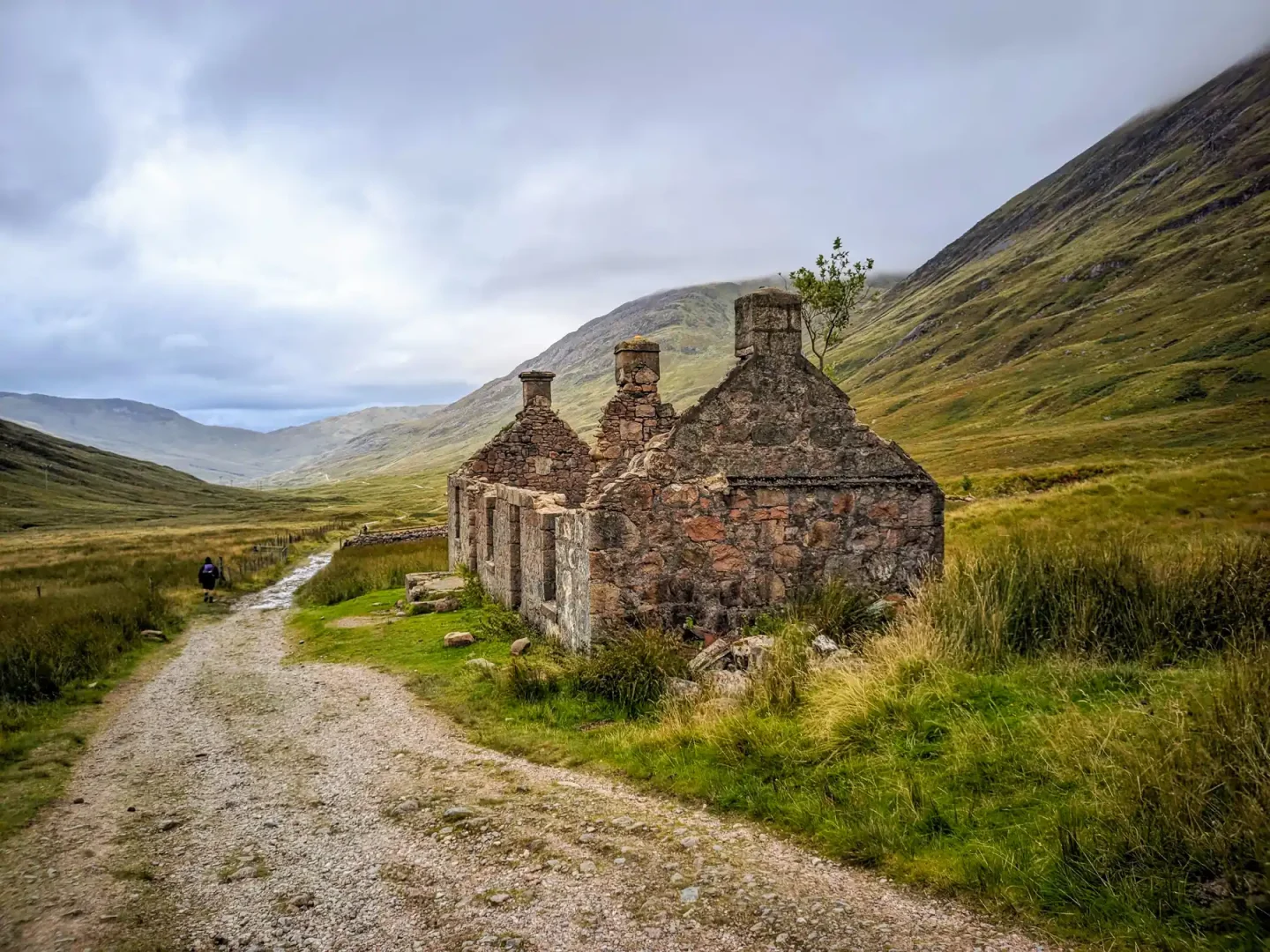 In a mountain landscape, a hiker passes a derelict stone building, with three chimneys but no roof, beside a broad stony track.