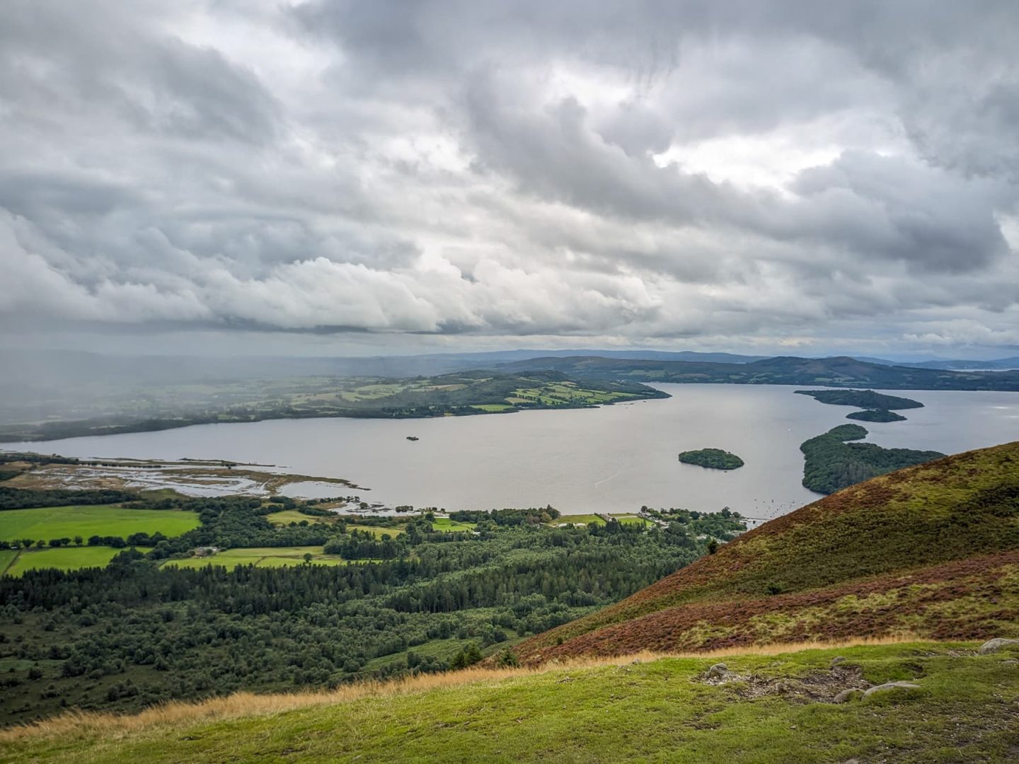 Looking down on Loch Lomond and some islands from Conic Hill. The lock is surrounded by hills and forests and low cloud is forming in the grey sky.