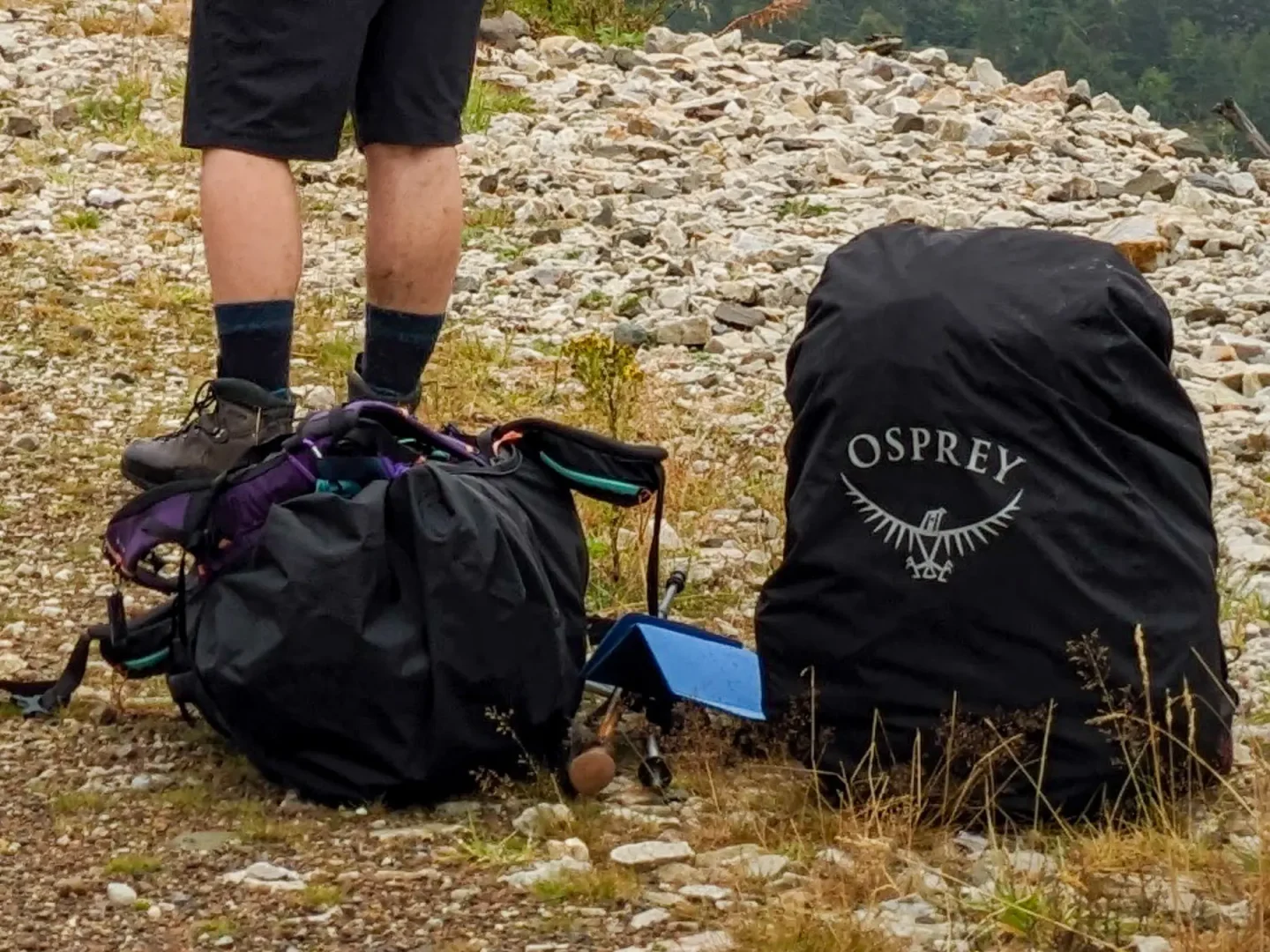 Two rucksacks on the ground. One is upright with a black cover and the Osprey brand logo and the other is lying. A pair of walking poles and a sit mat are between the two.