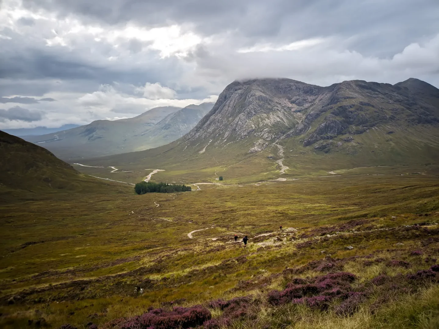 In the distance, hikers climb a steep grassy hill towards the top of a mountain. Behind them are the grand mountains of Glen Coe.
