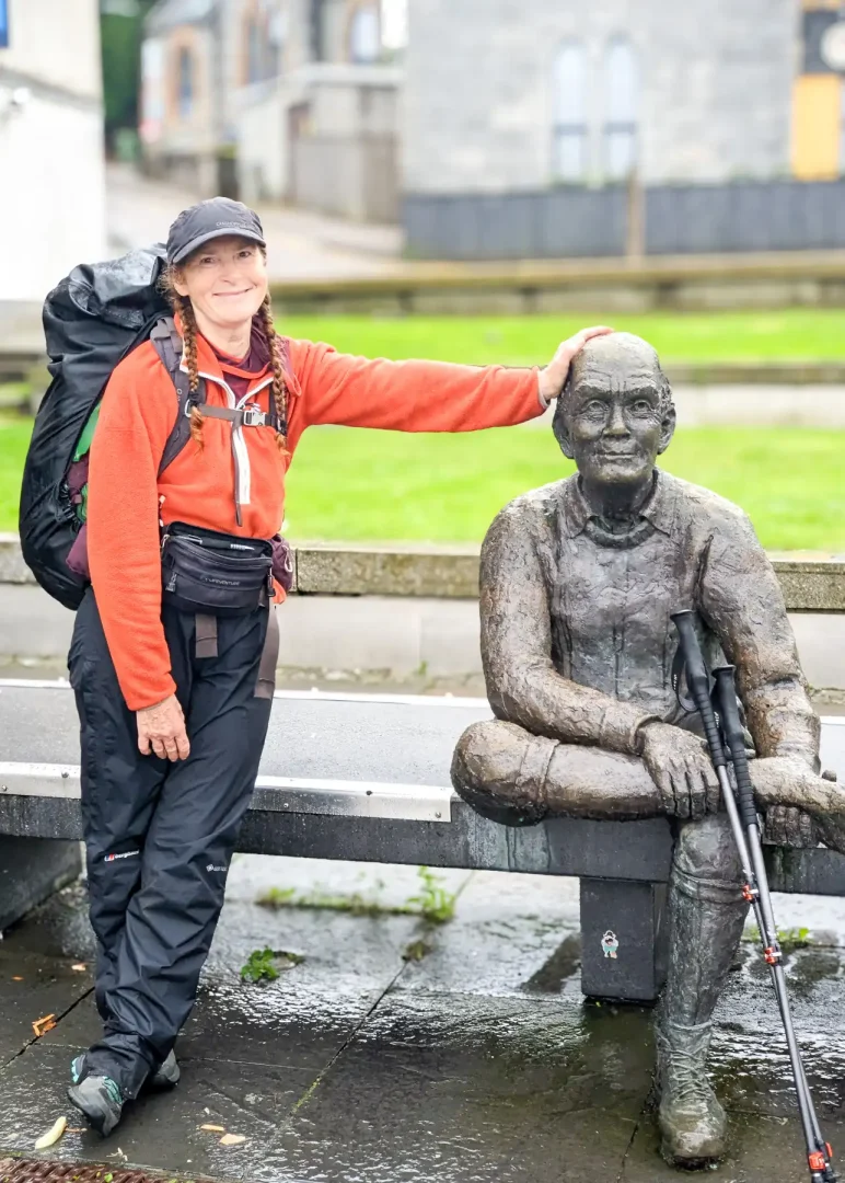 I'm smiling at the camera leaning on the sitting statue of The Walker, marking the end of the West Highland Way. My walking poles are leaning up against his leg, and of course, it's raining!