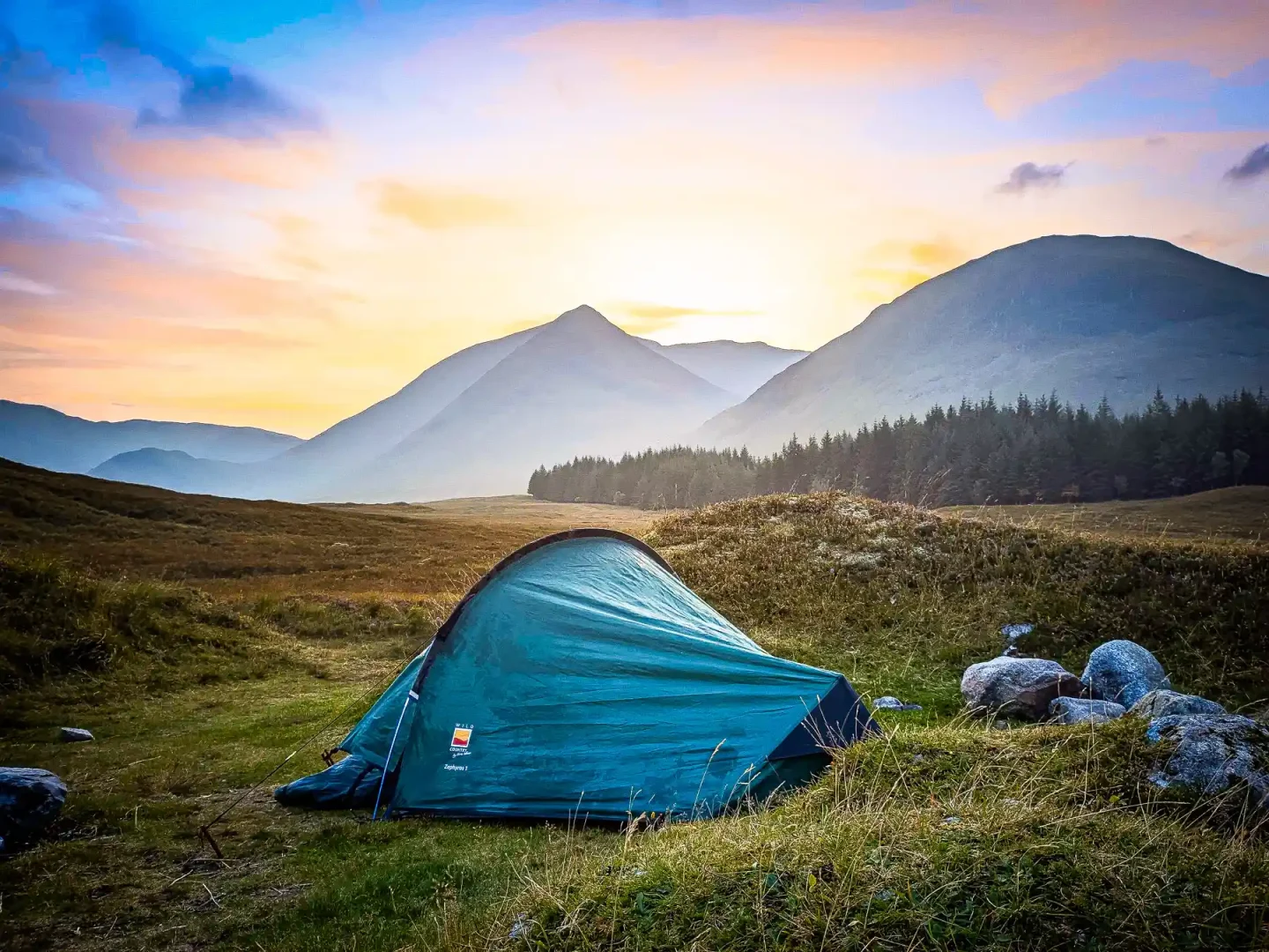 Wild camp on Rannoch Moor - there's a glorious sunset above the mountains, and my tent is pitched in the foreground, with a forest behind at the base of a mountain.