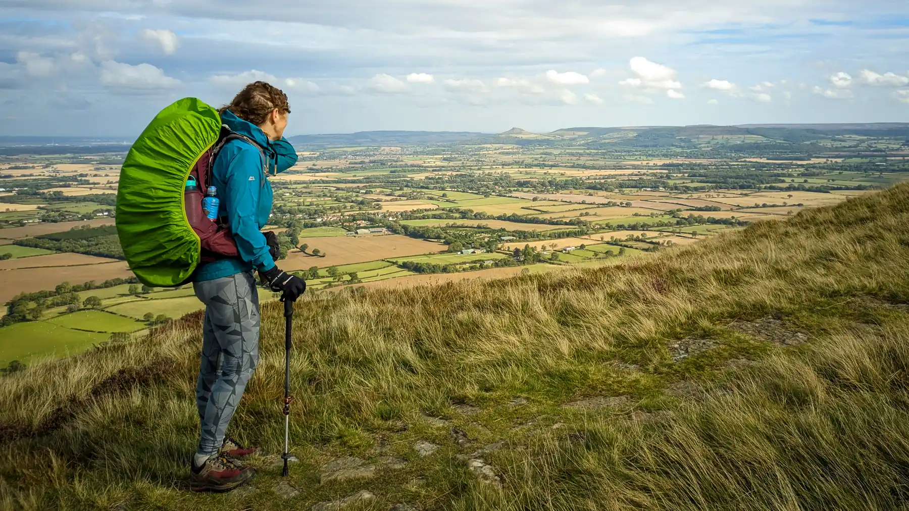 A hiker (Stephie) stands on top a of an escarpment looking out across fields to the horizon, where Roseberry Topping rises distinctly above the surrounding hills.