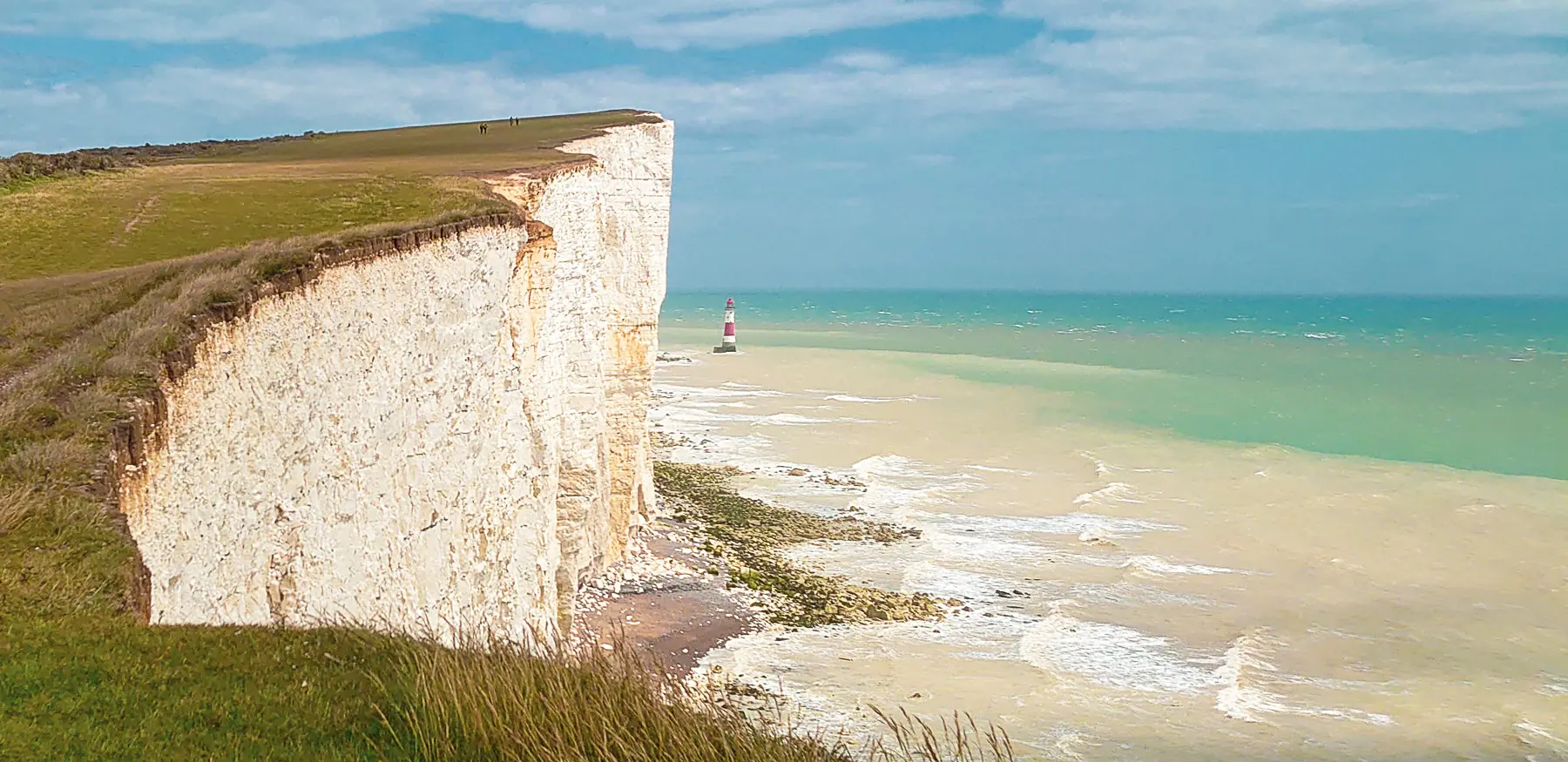 A huge white cliff towers above a red and white striped lighthouse far below on a sandy beach.