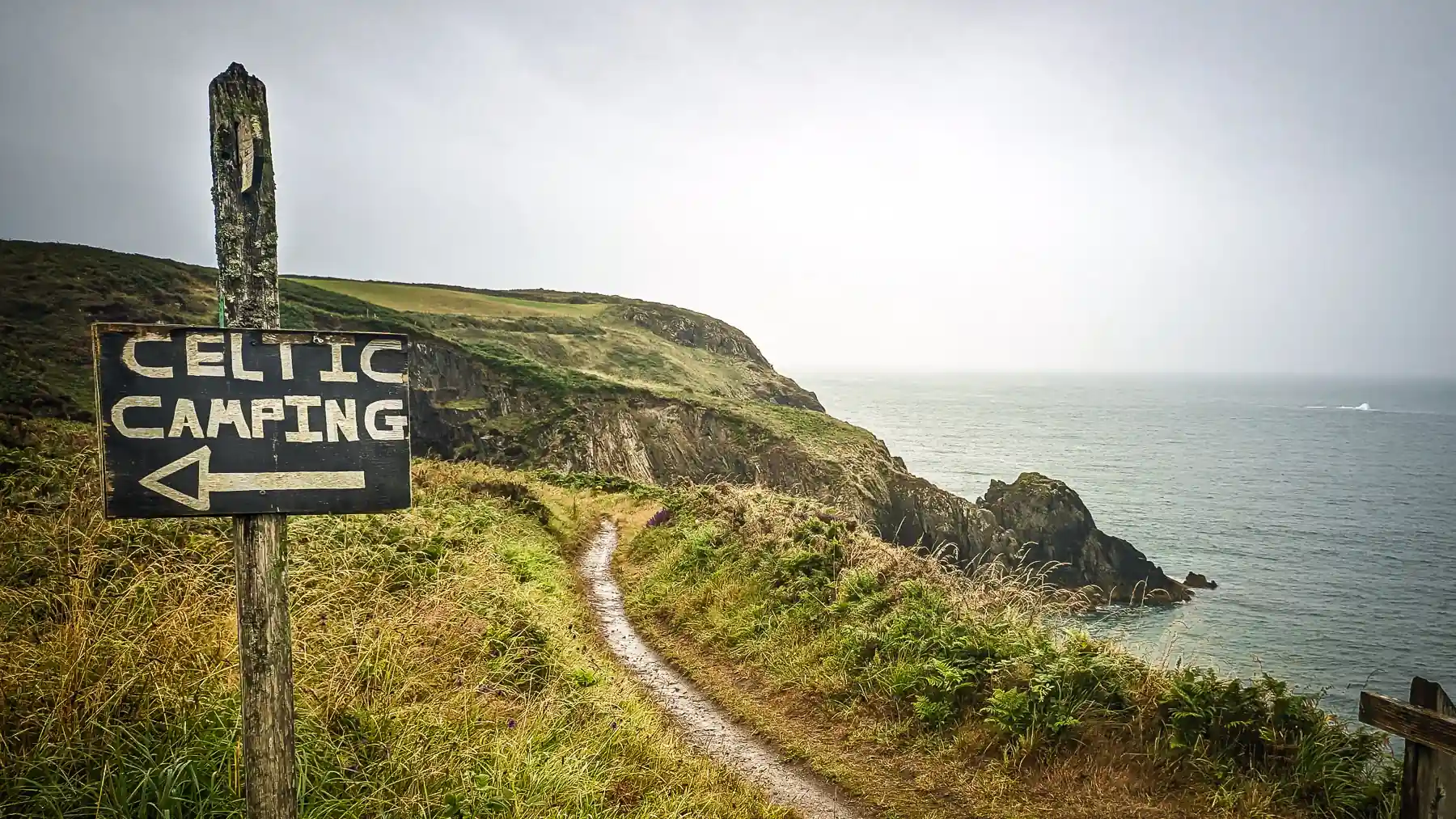A rough wooden signpost to 'Celtic Camping' is next to the cliff path on a rainy day