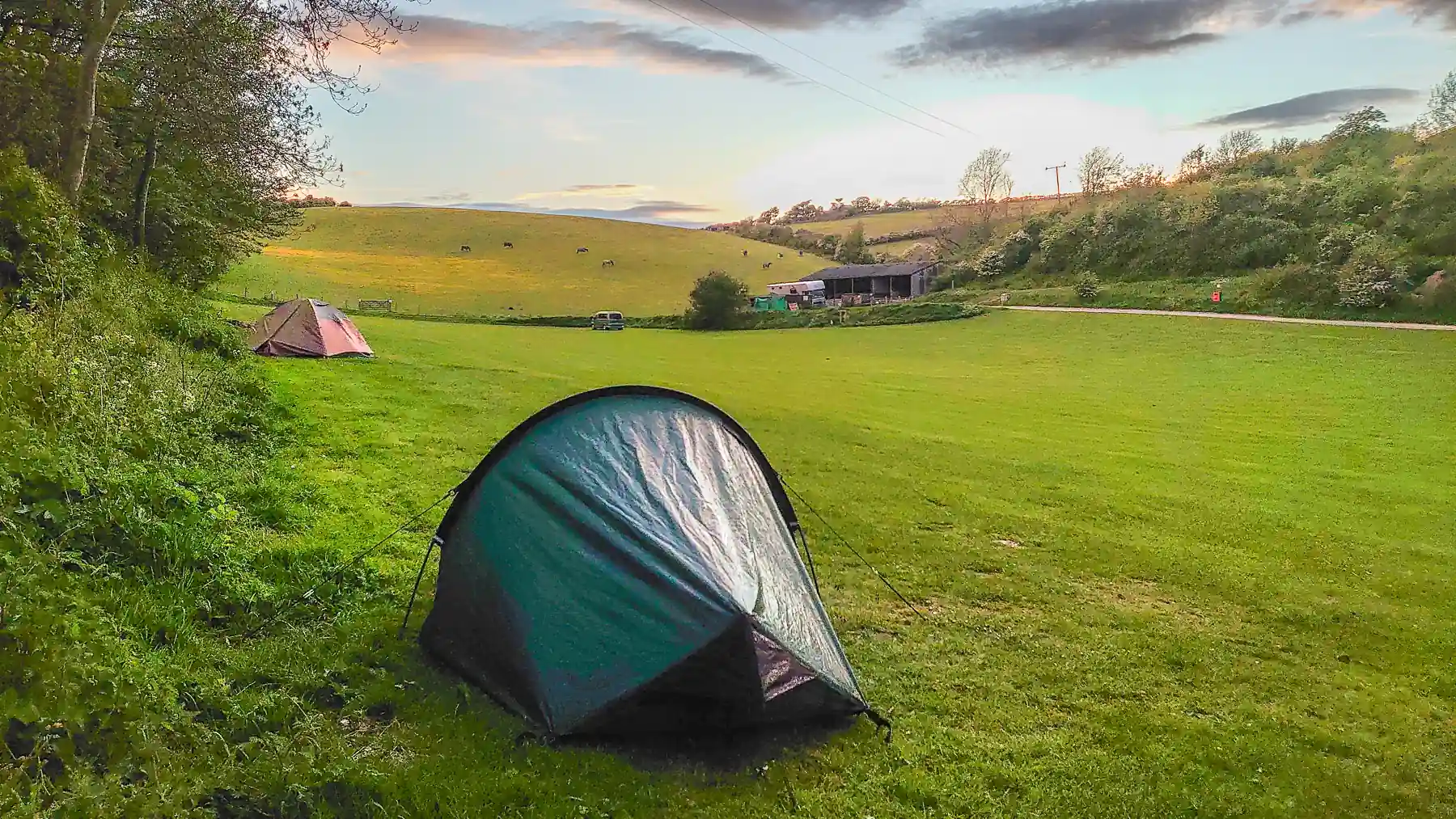 Two small tents are pitched close to a hedge on a large field. In the distance, there's a barn in a neighbouring field and rain clouds drift above.