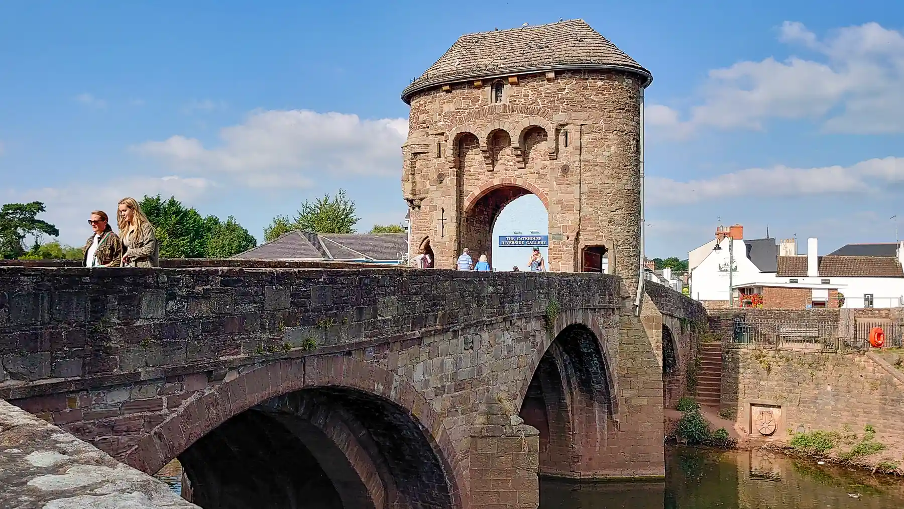 People are walking through a medieval stone gatehouse to cross an arched bridge over a river on a sunny summer's day.