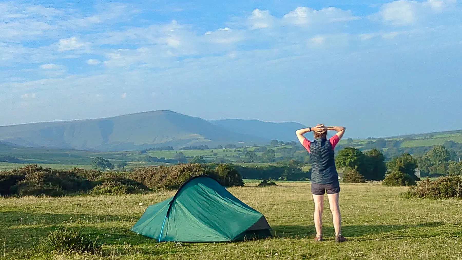 A woman stands beside a small backpacking tent looking out across fields to mountains on the horizon.