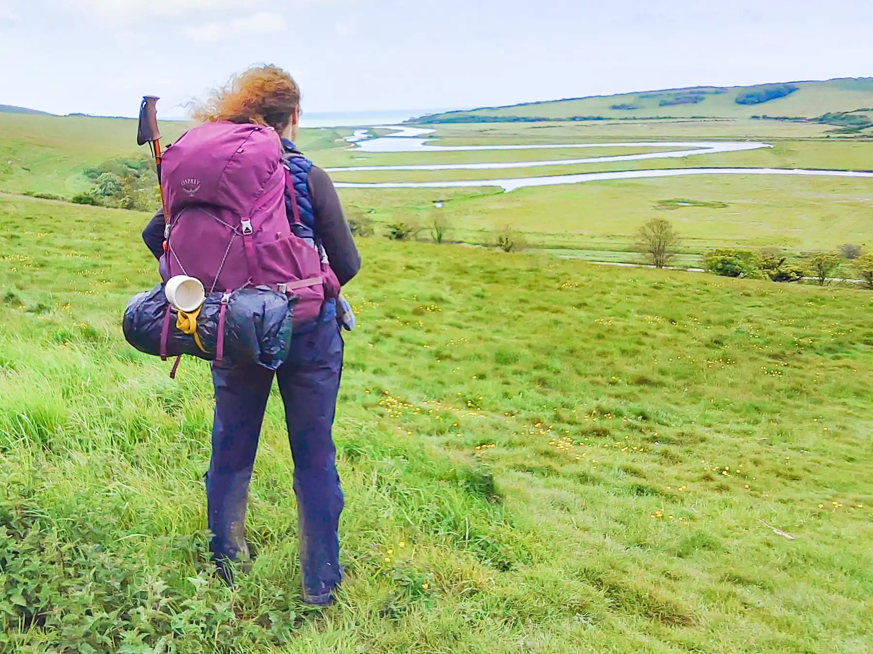 A hiker stands looking out along a winding river towards the sea. She wears a heavy rucksack with a tent and mug attached to the pack.