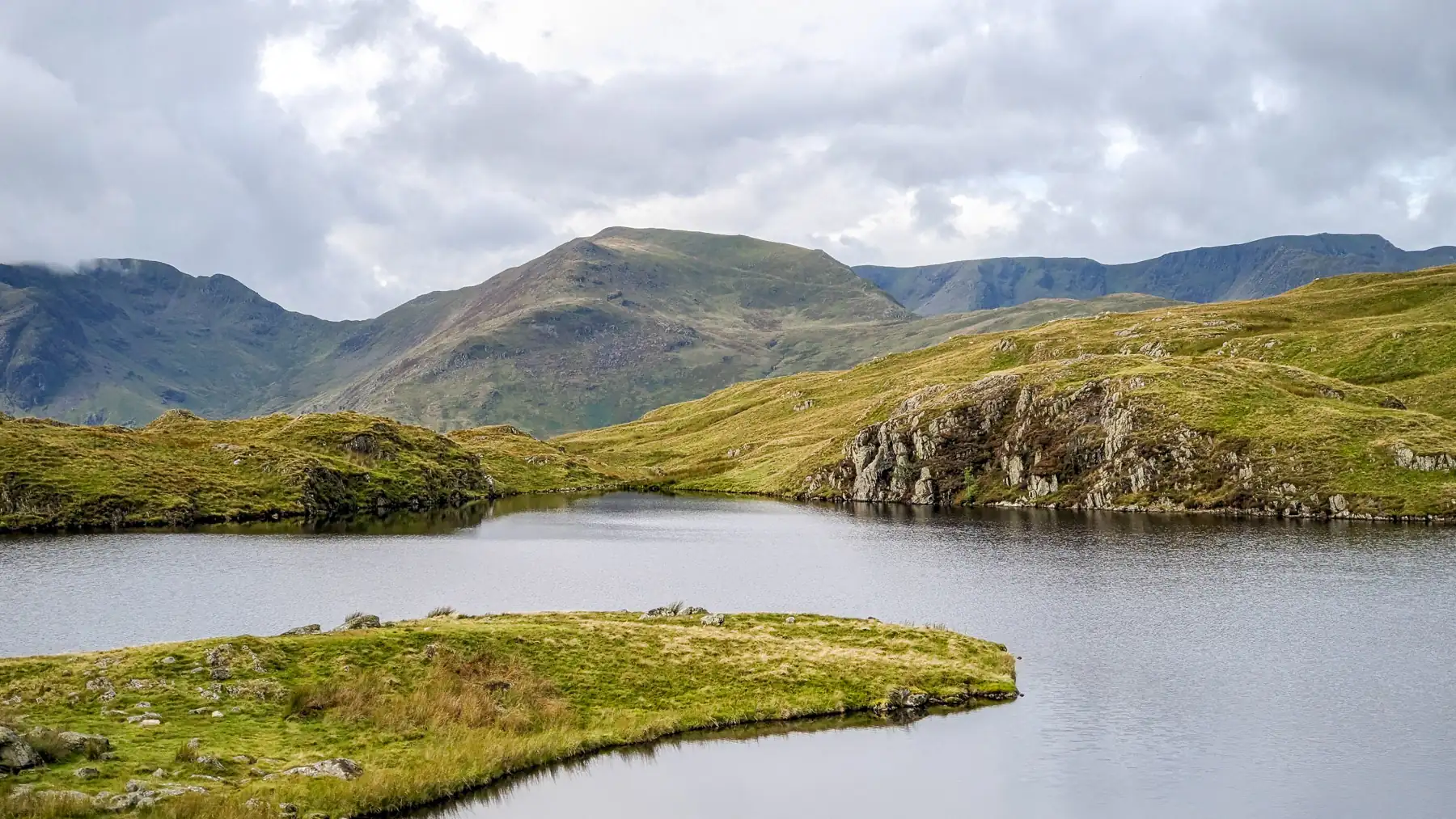 Angle Tarn in the Lake District - a small lake with a finger of land stretching into it from the foreground, with mountains behind, on a cloudy, grey day.