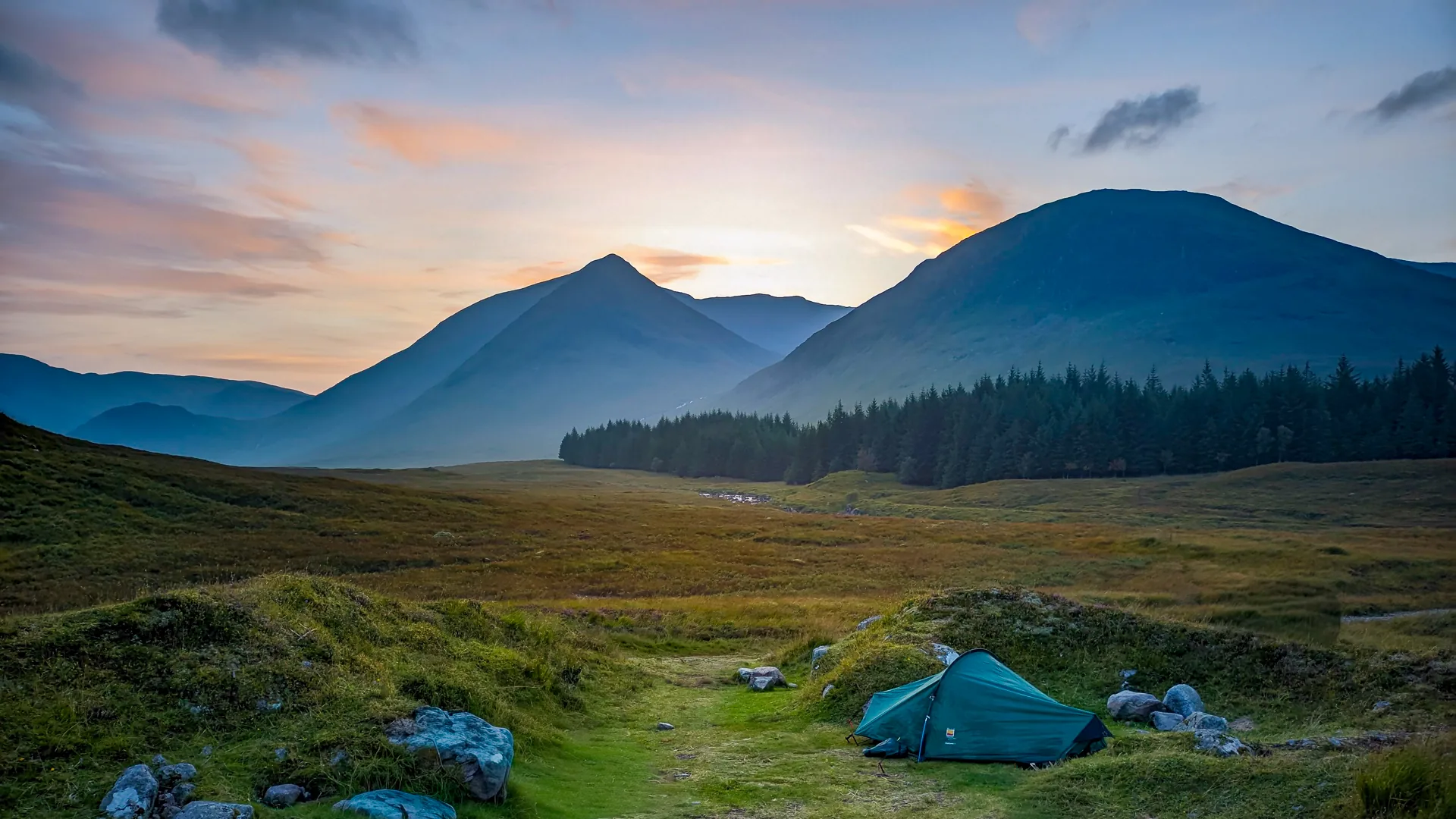 A backpacking tent is nestled beside a hump of grass on Rannoch Moor. A pine forest and tall mountains are on the near horizon, silhouetted by a dusky sunset.