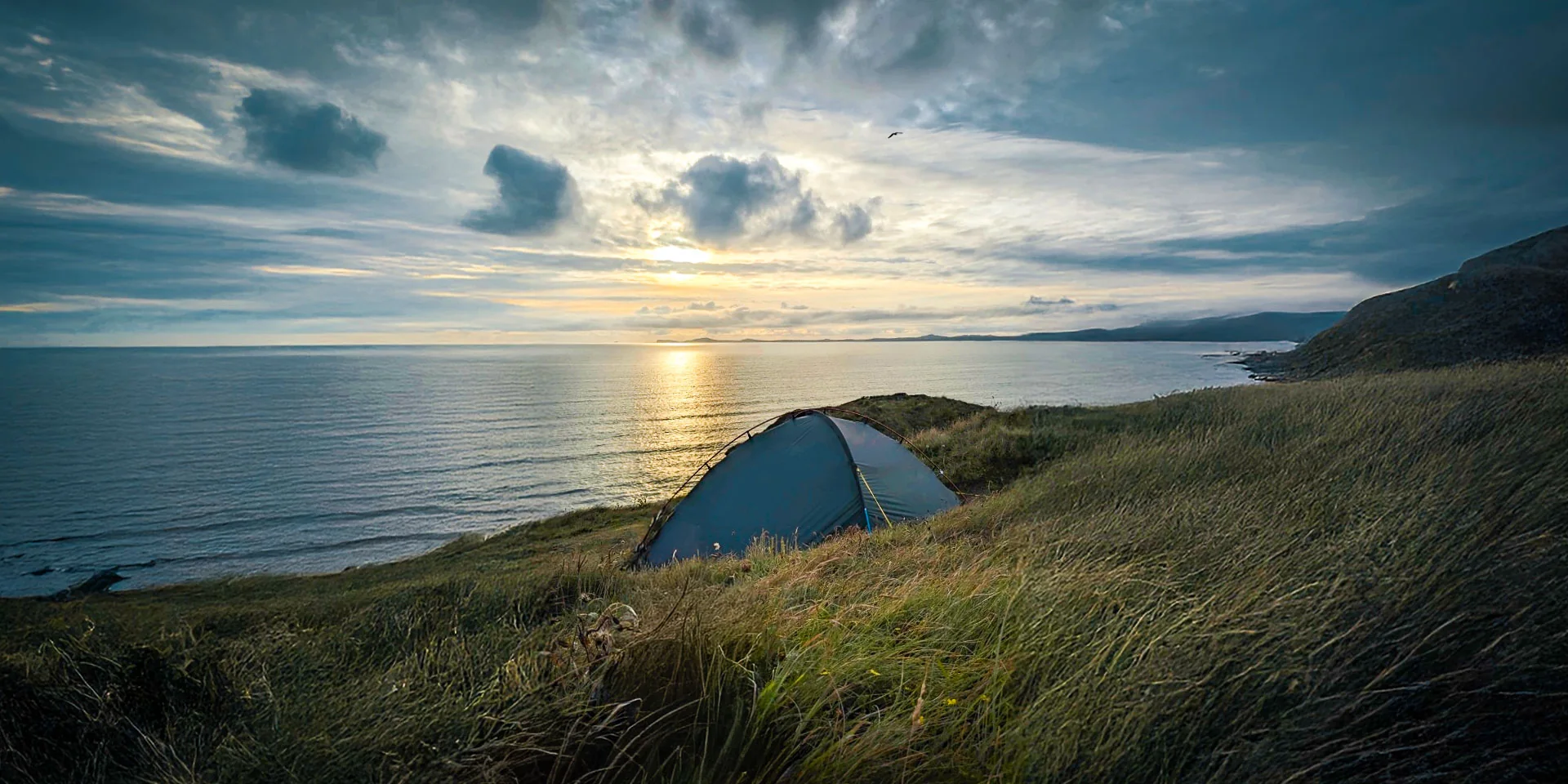 A tent is pitched on a grassy cliff-edge overlooking a sunset over the sea.