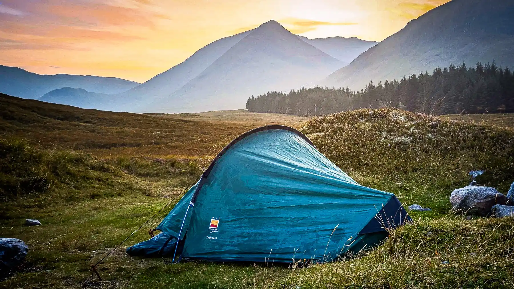 A backpacking tent is pitched up at dusk at the bottom of mountains in the Scottish Highlands. The sky is an array of soft oranges and pinks.