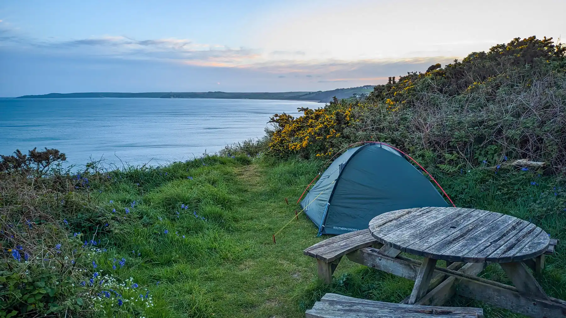 A tent is pitched on the cliffs beside a rickety picnic bench. The sun has set and wispy yellow clouds pass above the calm sea