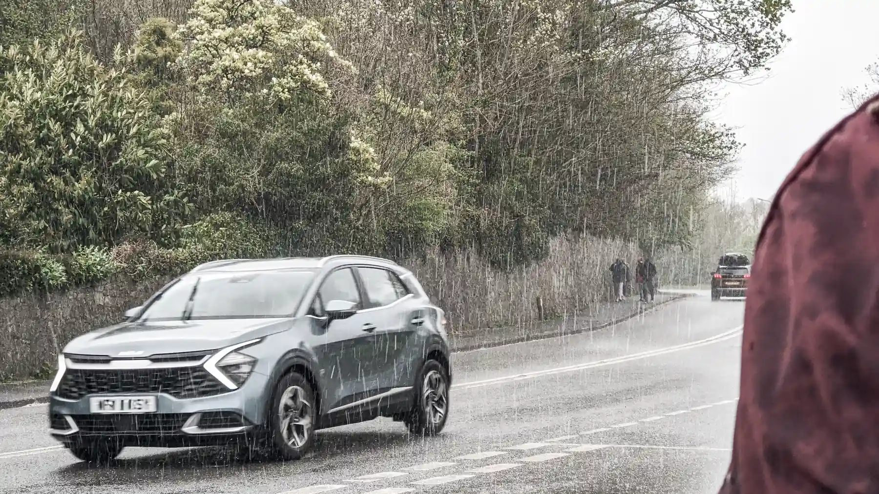 Pedestrians walk by and a car drives past in a very heavy hailstorm. My rucksack stays dry in a bus stop shelter. The photo has been edited with a grey, washed-out feel that highlights the cold and miserable atmosphere.