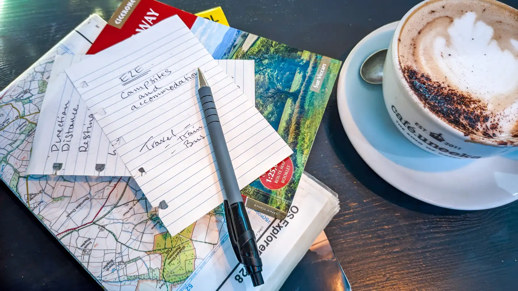 A map, guide and paper notes and pen are in a stack beside a cappuccino in a cafe