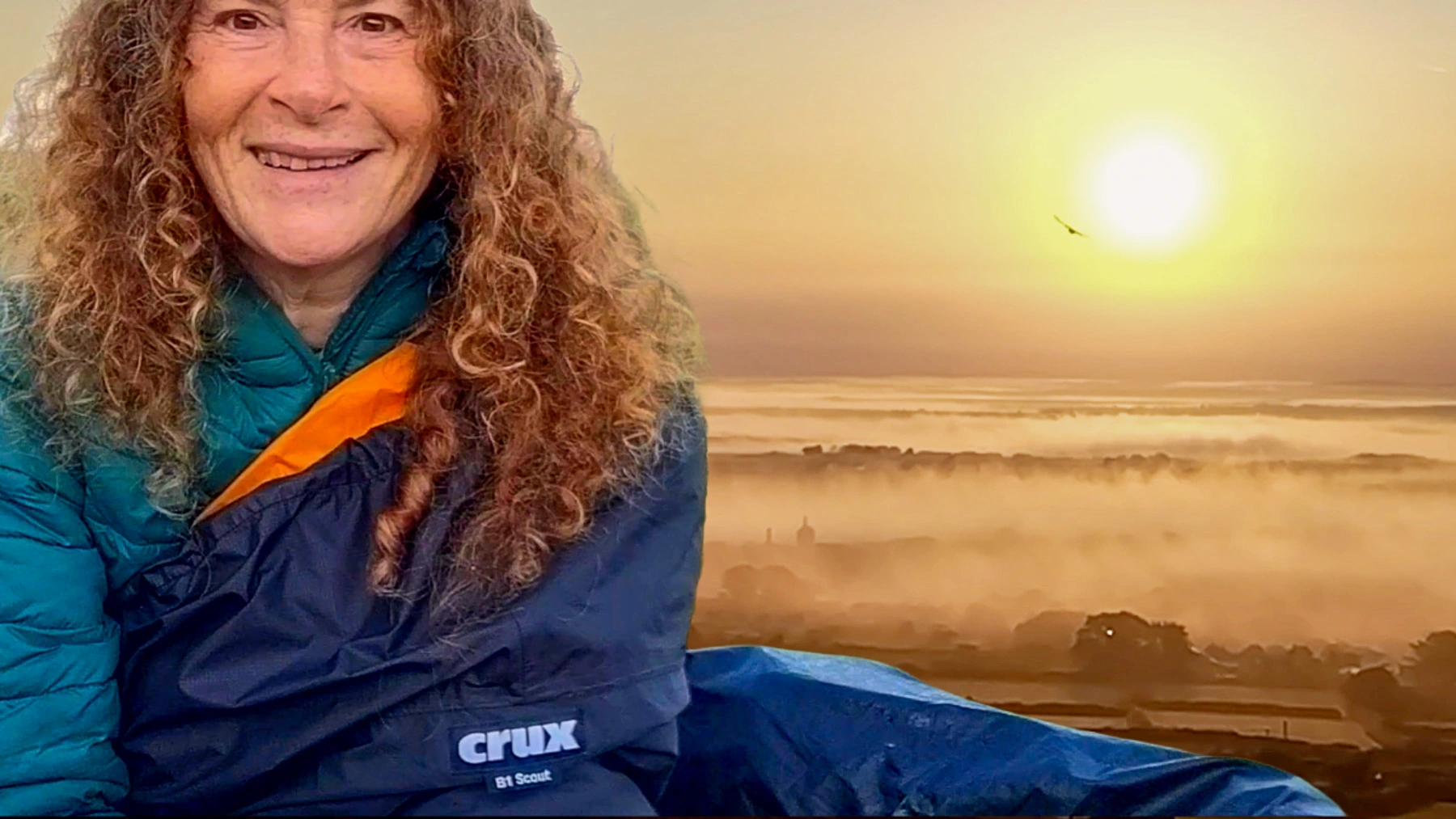 I'm sat up in a bivvy bag smiling at the camera. There's a misty sunrise behind.