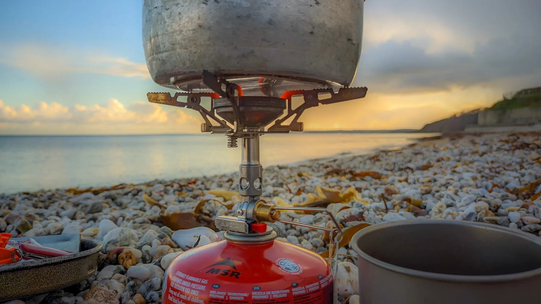 A view of a beach beyond a camping gas stove. There's a pot on the stove and a mug waiting beside it.