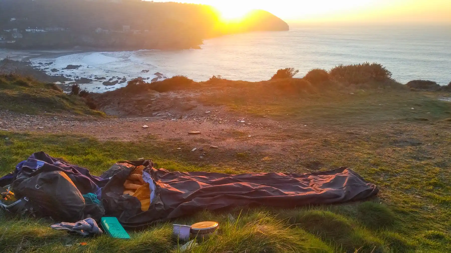 A bivvy bag and rucksack lie on the grass on a clifftop. The sun is setting, dipping below a headland in the distance, giving the scene an orange glow.