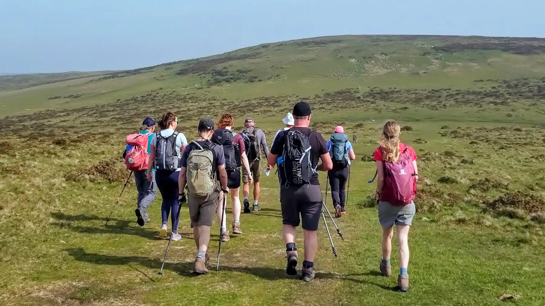 A group of hikers walking in the hills are seen from behind. They're all carrying daysacks, some are using walking poles, and they're wearing an array of clothes suitable for a day hike.