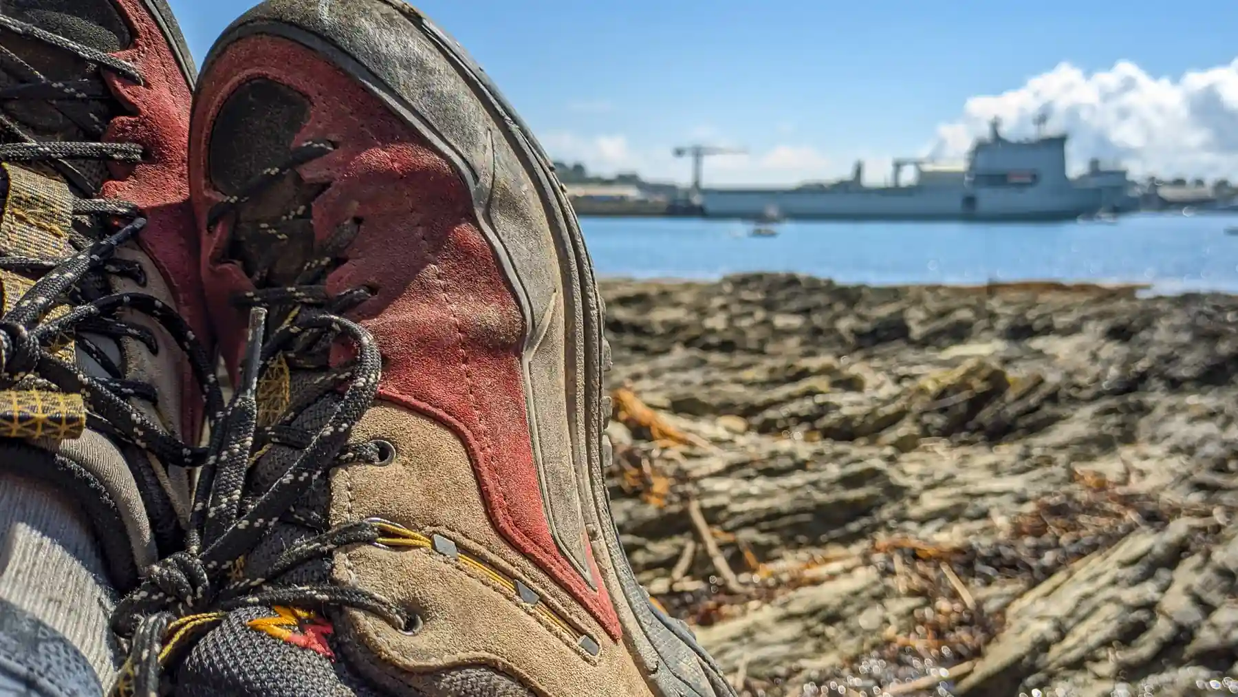 A close up of walking shoes - resting on a rocky beach with the sea and a ship in the background