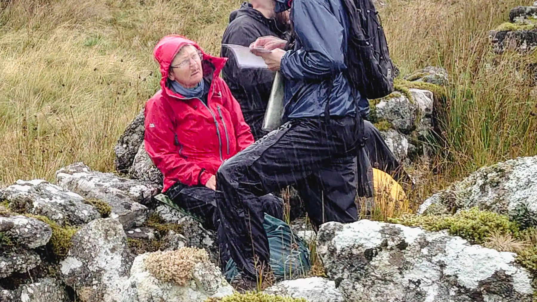 An instructor in a red jacket sits on a rock talking to participant who's reading a map. Everyone is soaking wet in torrential rain on Dartmoor.