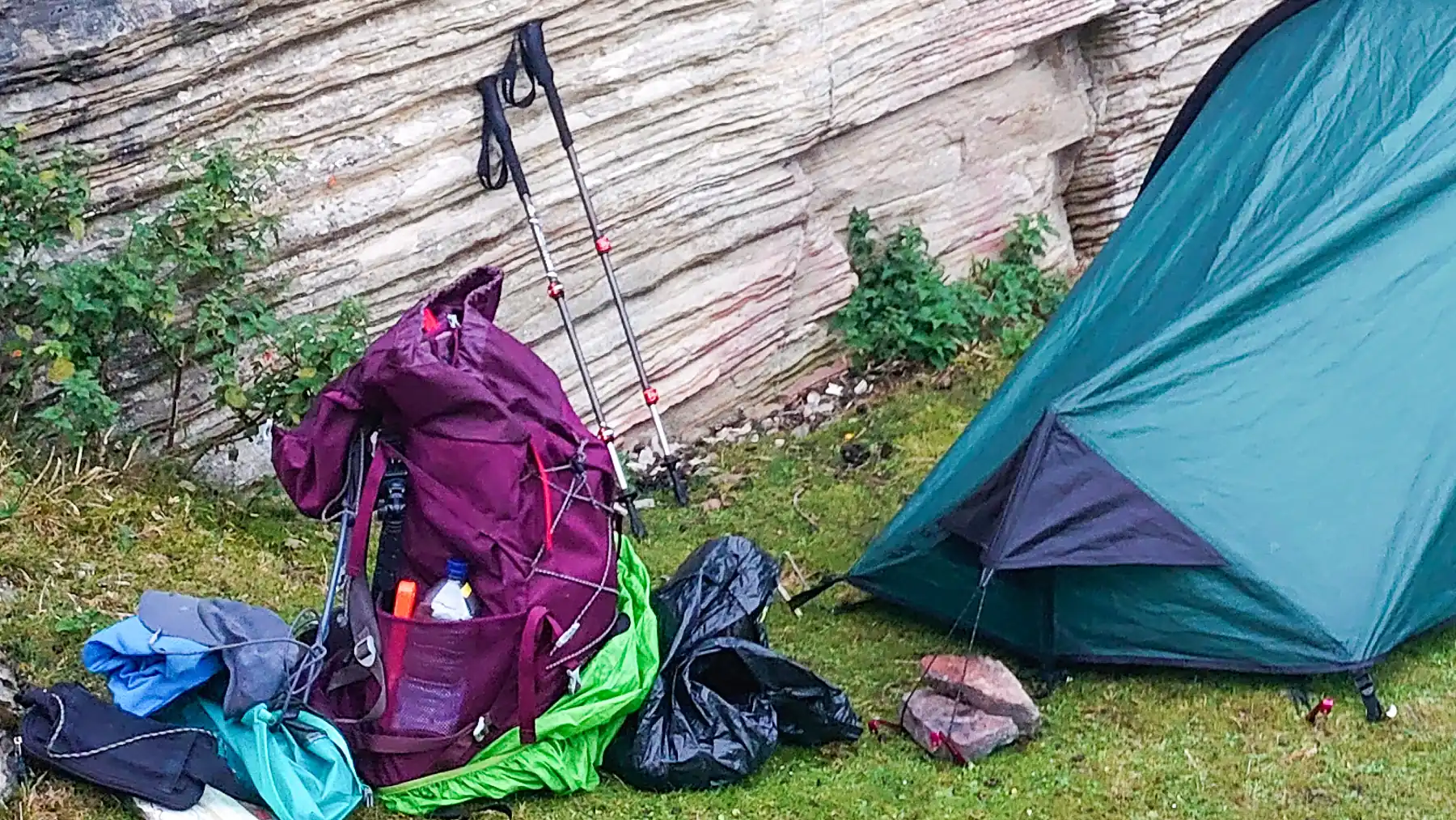Wild camping beside a cliff wall with an open rucksack and the contents spread on the grass around it.